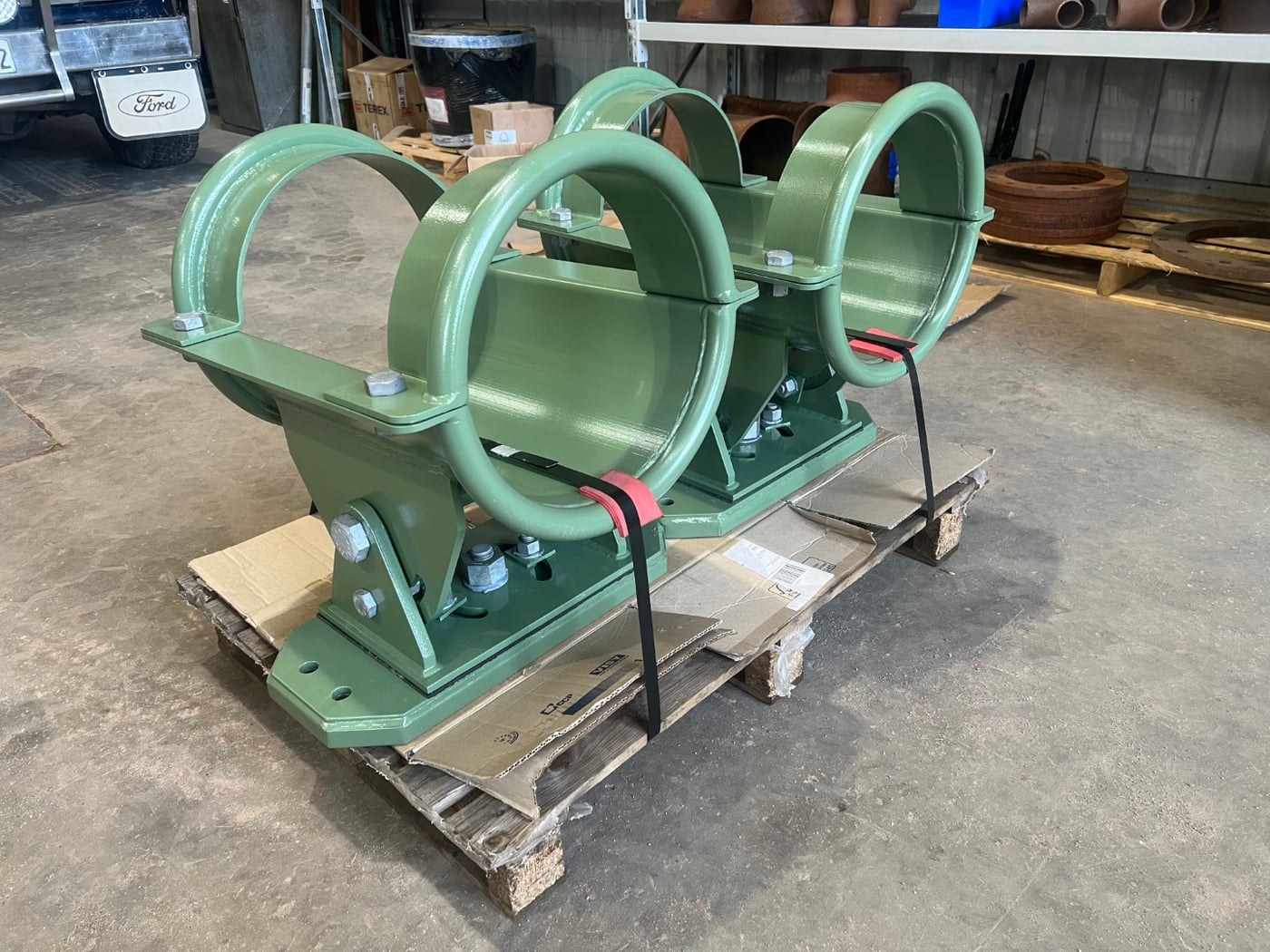 Pair of green painted fabricated pipe support clamps on a pallet in the workshop